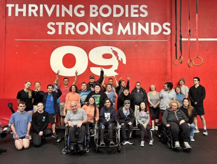 A large, diverse group of people pose together in an indoor CrossFit gym against a bright red wall.