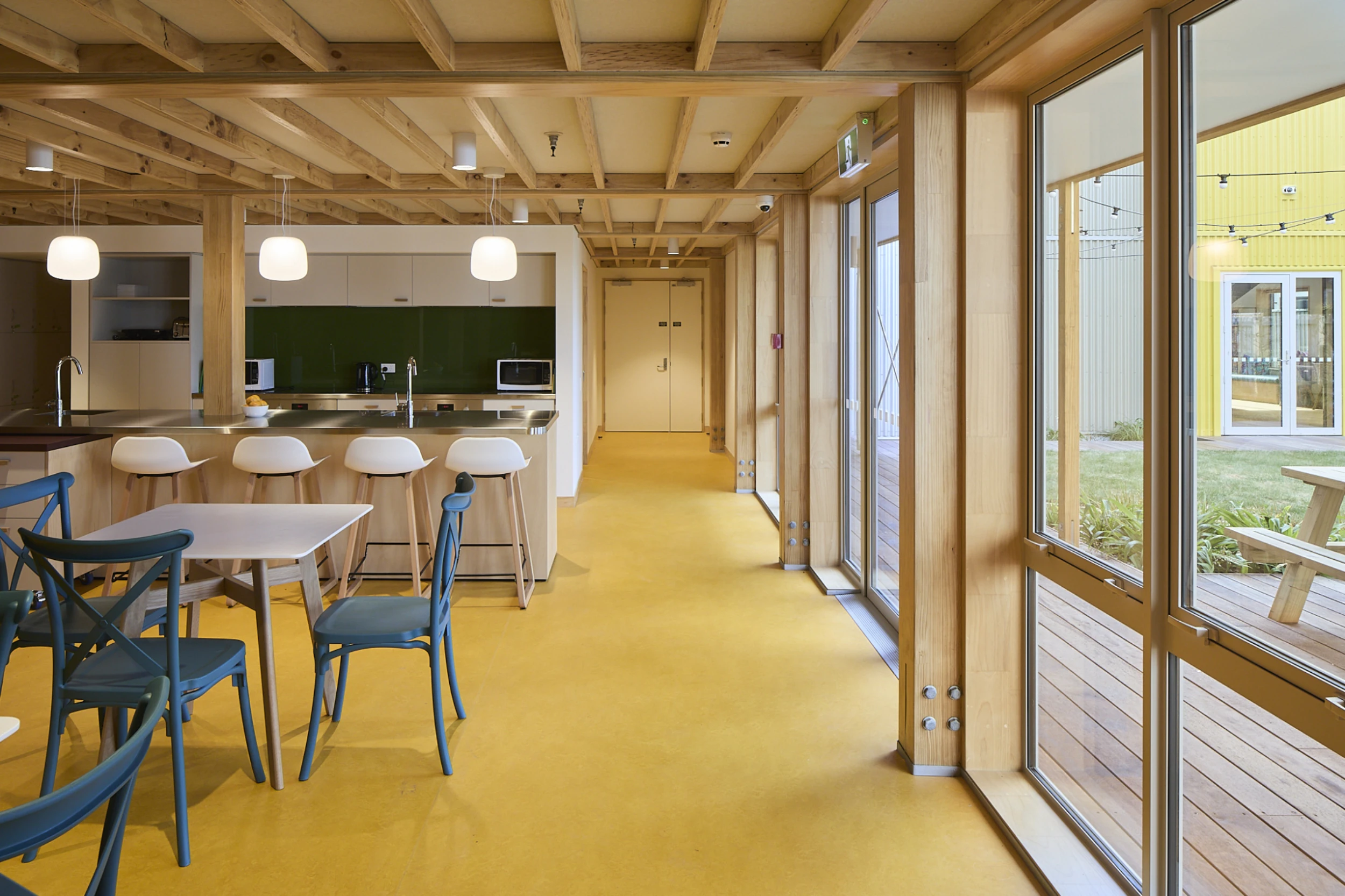 Bright, modern kitchen with yellow floors, wooden ceiling beams, and large windows. White chairs and blue chairs around a dining table. Warm, inviting tone.