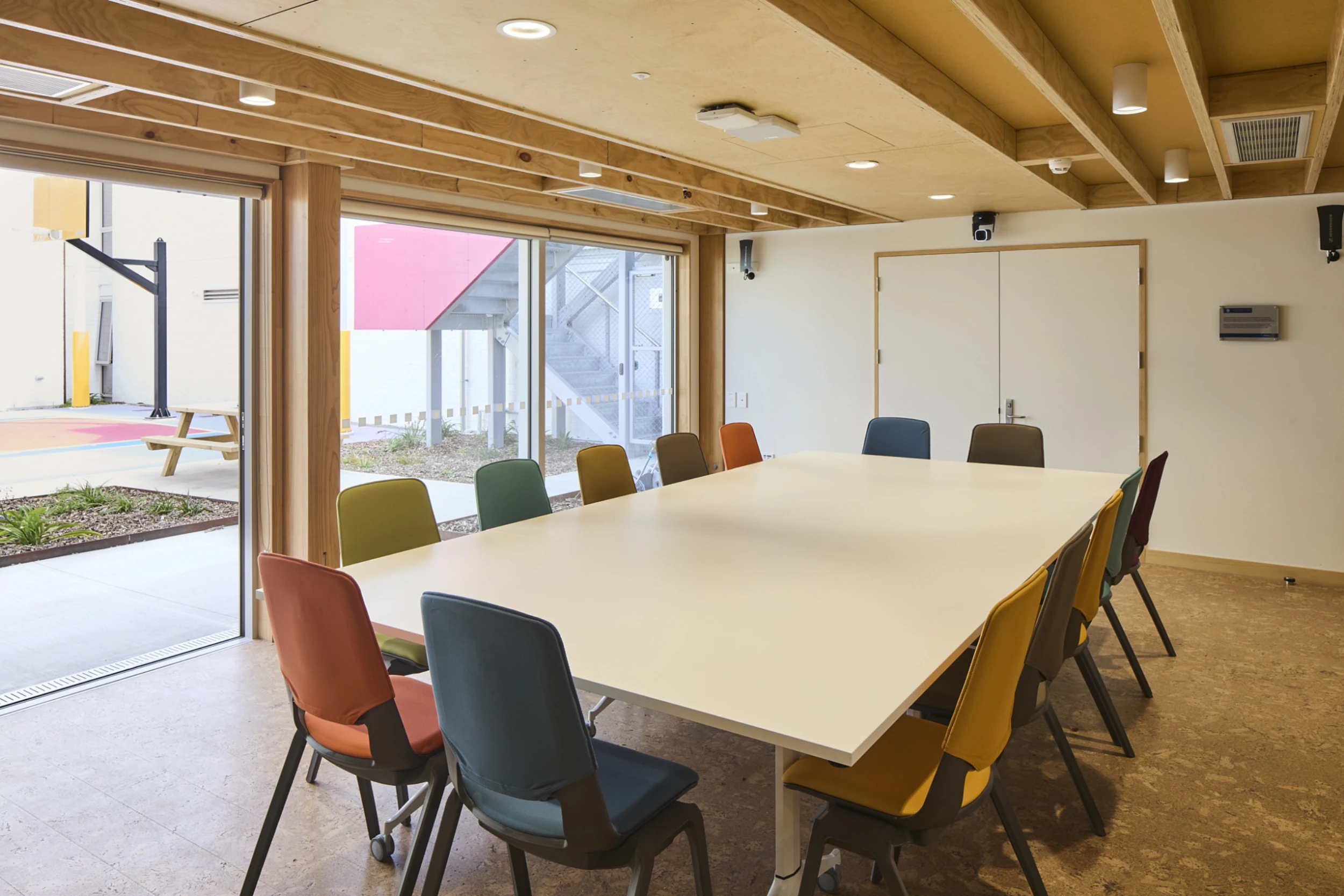 A modern meeting room with a long white table and ten colorful chairs. Large windows reveal an outdoor area with a picnic table, creating an open and inviting atmosphere.