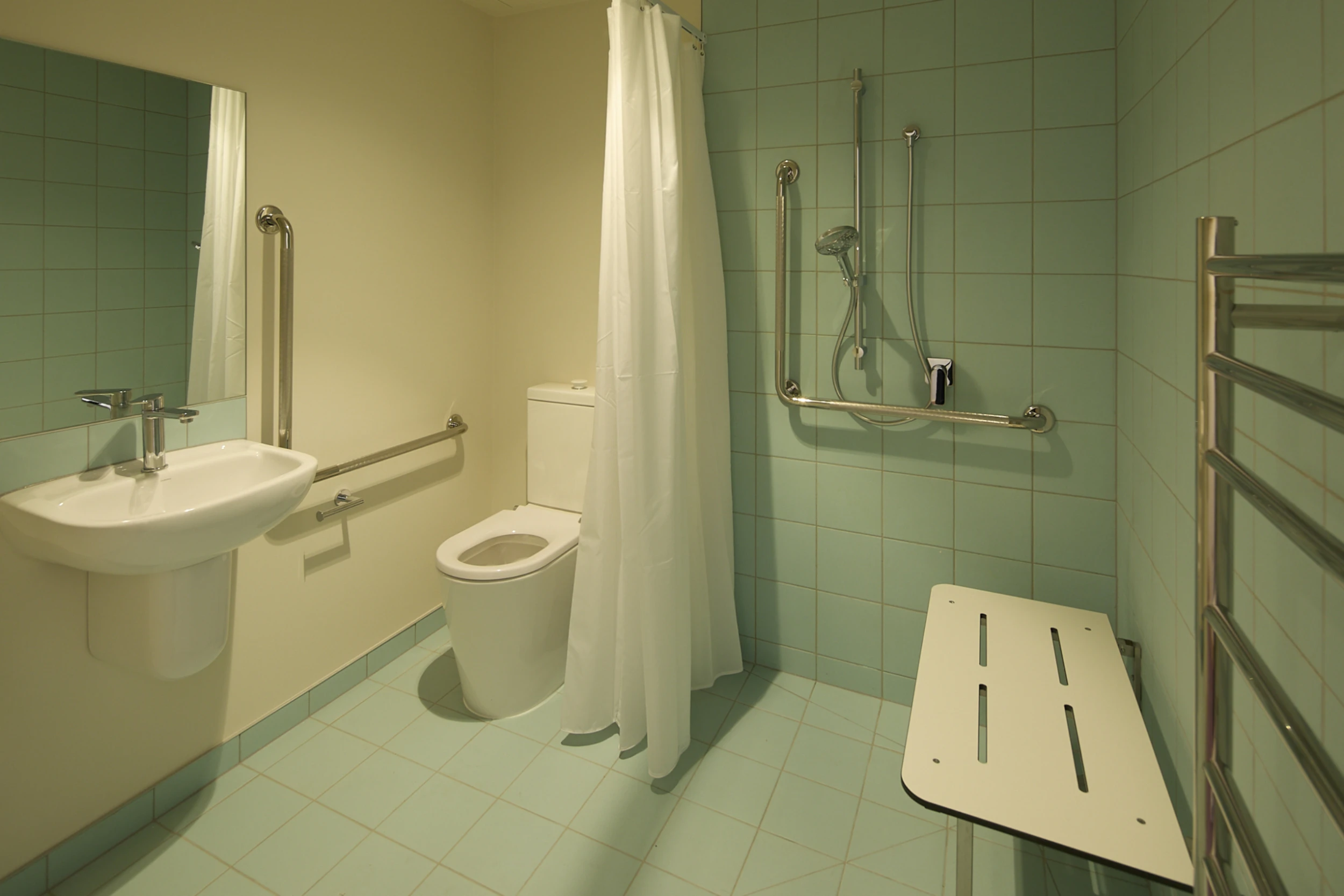Accessible bathroom with blue tiles, featuring a toilet, sink, and shower with grab bars, a curtain, and a foldable bench. Clean, functional design.