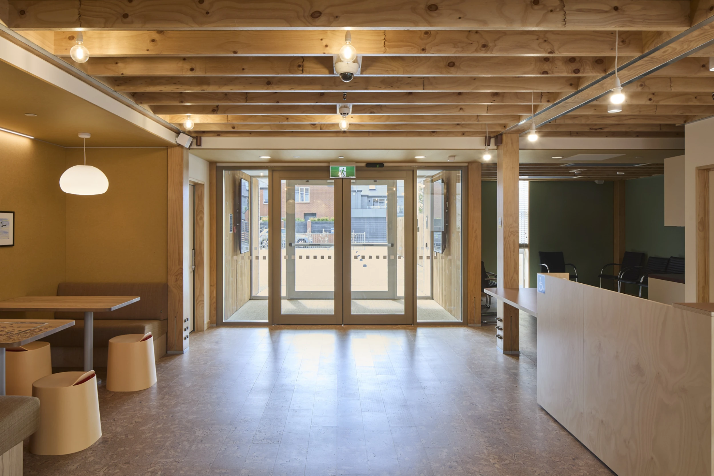 Spacious room with wooden ceiling beams and floors, large glass doors at the back. Cozy booth with modern stools on the left, and a reception desk on the right.