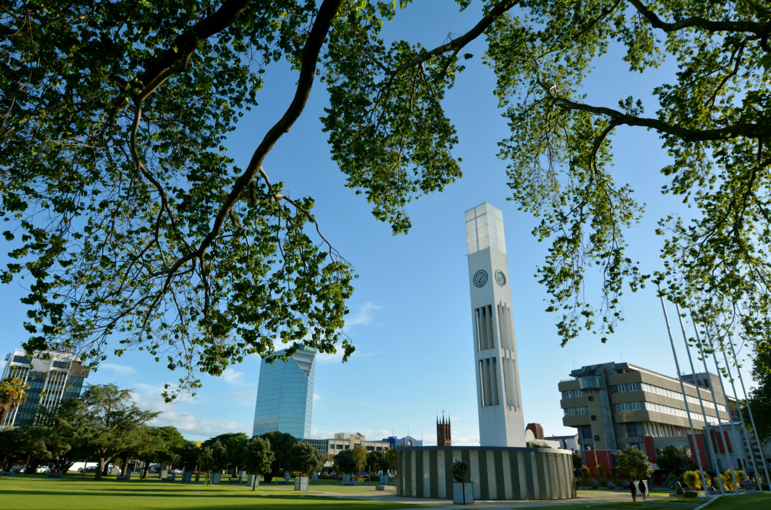 Tall white clock tower in a sunny urban park. Green branches frame the scene, with modern buildings and clear blue sky in the background. Peaceful atmosphere.