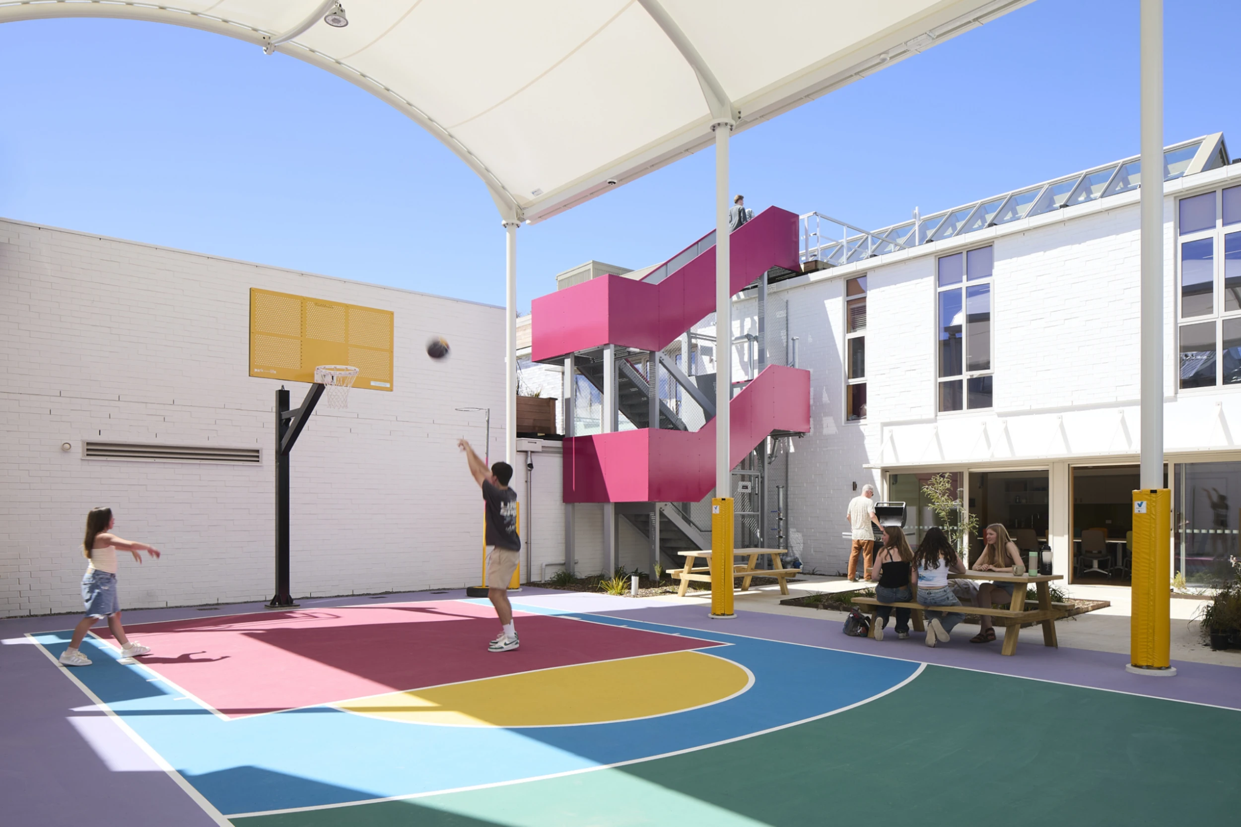 A vibrant outdoor basketball court with colorful markings in red, yellow, and blue. A young man and girl play basketball under a white canopy. In the background, a pink staircase is attached to a white building, with people sitting at picnic tables nearby, creating a lively and playful atmosphere.