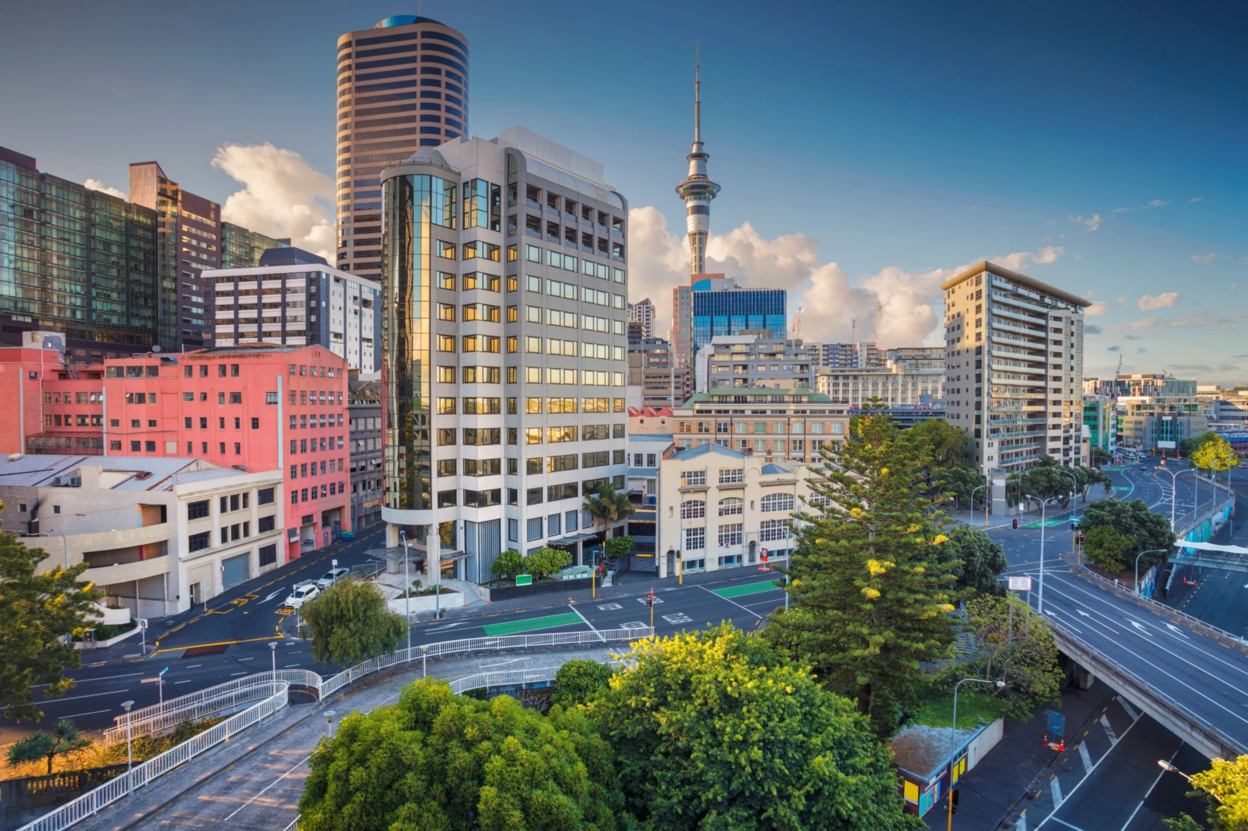 A vibrant cityscape of Auckland, featuring modern high-rises, the iconic Sky Tower, and lush greenery, under a clear blue sky at dusk.