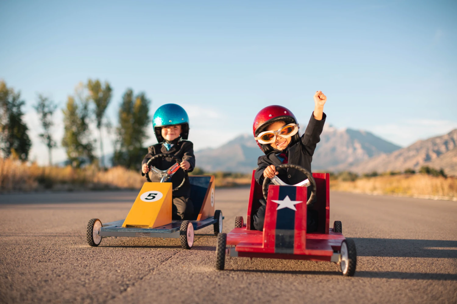 Two children in red and blue helmets race in homemade go-karts on a sunny day. The child in the red kart celebrates with a raised arm, conveying excitement. Mountains and trees form the backdrop.