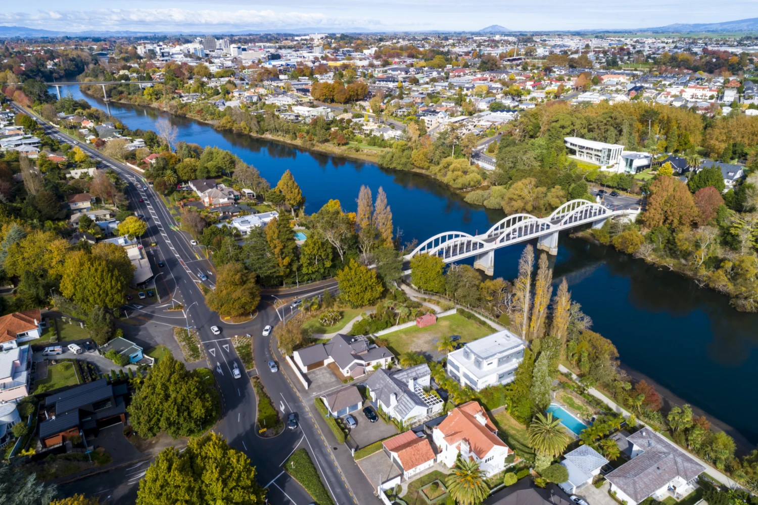 Aerial view of the serene Waikato River with an arched white bridge, surrounded by lush trees and a suburban neighbourhood of Hamilton. There is a clear blue sky and a distant cityscape.
