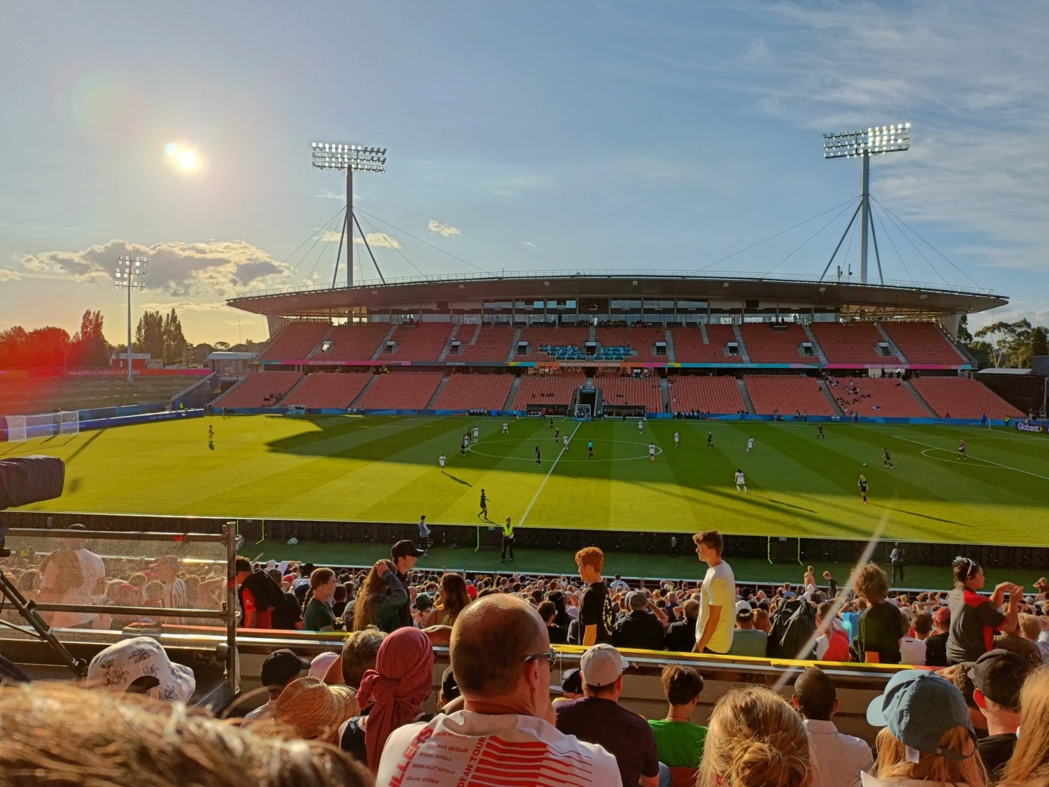 A vibrant rugby match unfolds at a sunlit FMG Stadium in Hamilton with a large crowd. The lush green field contrasts with the orange seats under clear skies.