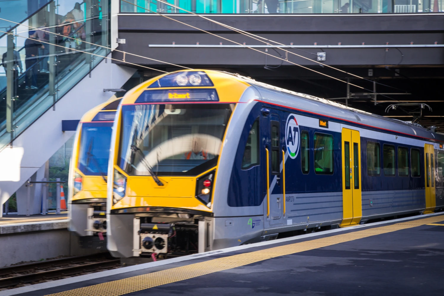 Two modern yellow and blue Auckland Transport passenger trains at a station platform, under a glass pedestrian bridge. Clear day, conveying a sense of urban transit efficiency.