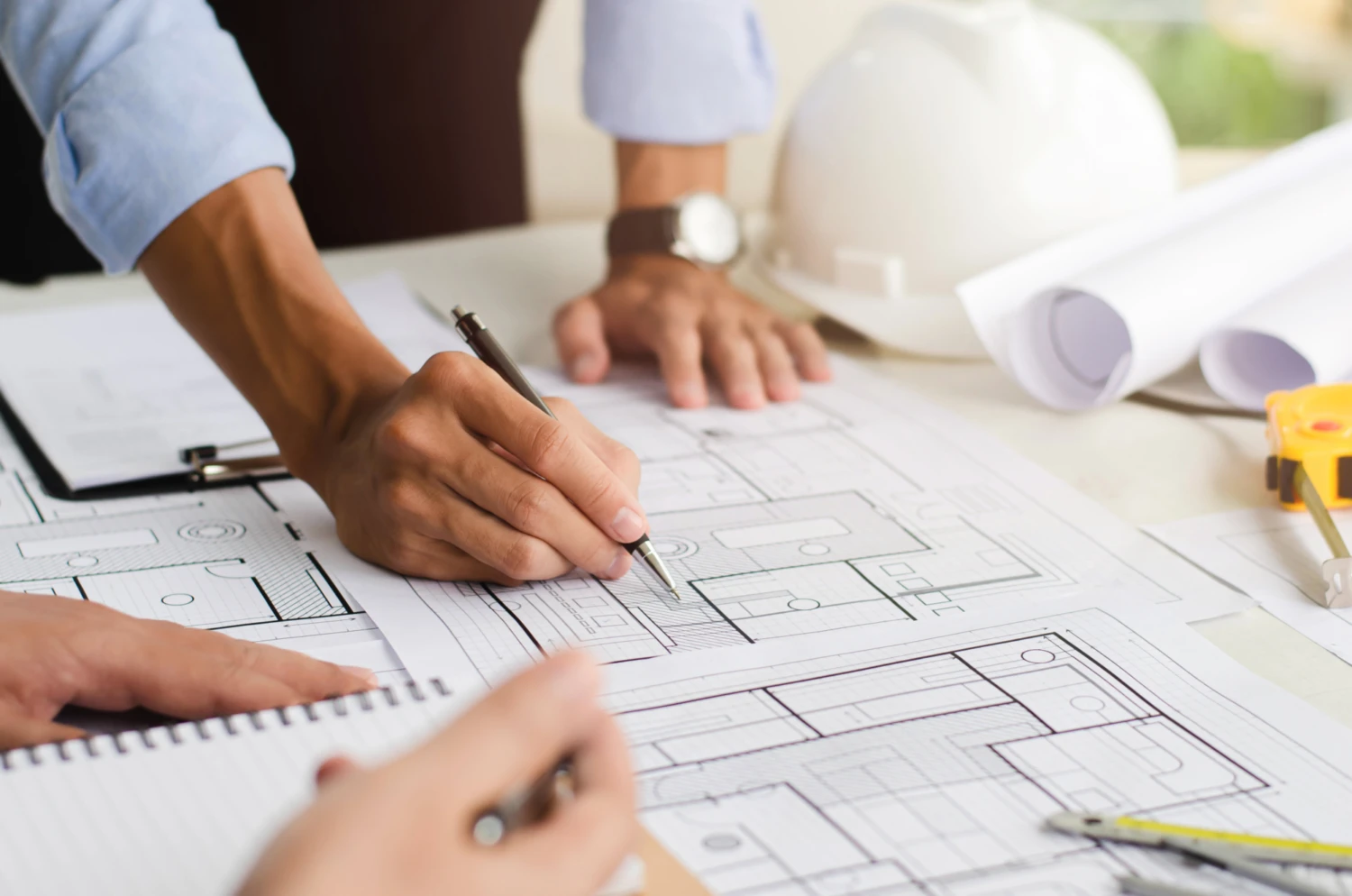 Hands of two people working on architectural blueprints at a desk, with a pencil, notepad, hard hat, and rolled plans, conveying teamwork and focus.