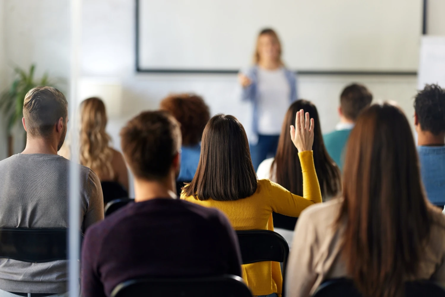 Audience seated indoors faces a presenter speaking at the front. A woman in yellow raises her hand, showing engagement. The setting is focused and educational.